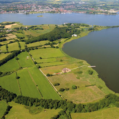 Luftbildaufnahme von Haithabu mit umgebendem Halbkreiswall, rekonstruierten Wikingerhäusern und Landebrücke am Haddebyer Noor sowie dem Ostseearm Schlei und die Stadt Schleswig im Hintergrund (2009)