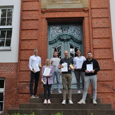 Die Absolvent*innen stehen vor dem Kreishaus und halten ihre Urkunden hoch