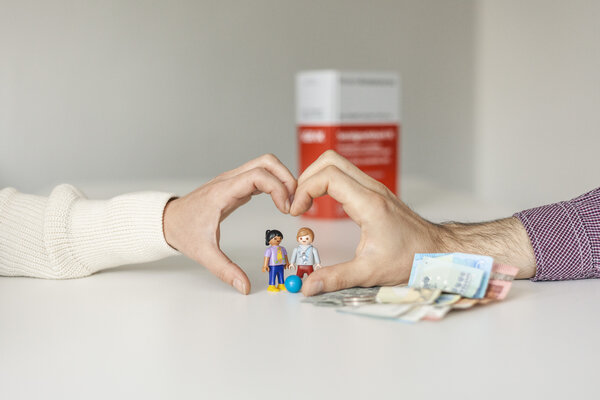 Zwei H&auml;nde formen ein Herz. In der Mitte davon stehen zwei Playmobil-Figuren. Daneben liegen Geldscheine und im Hintergrund steht ein Buch.